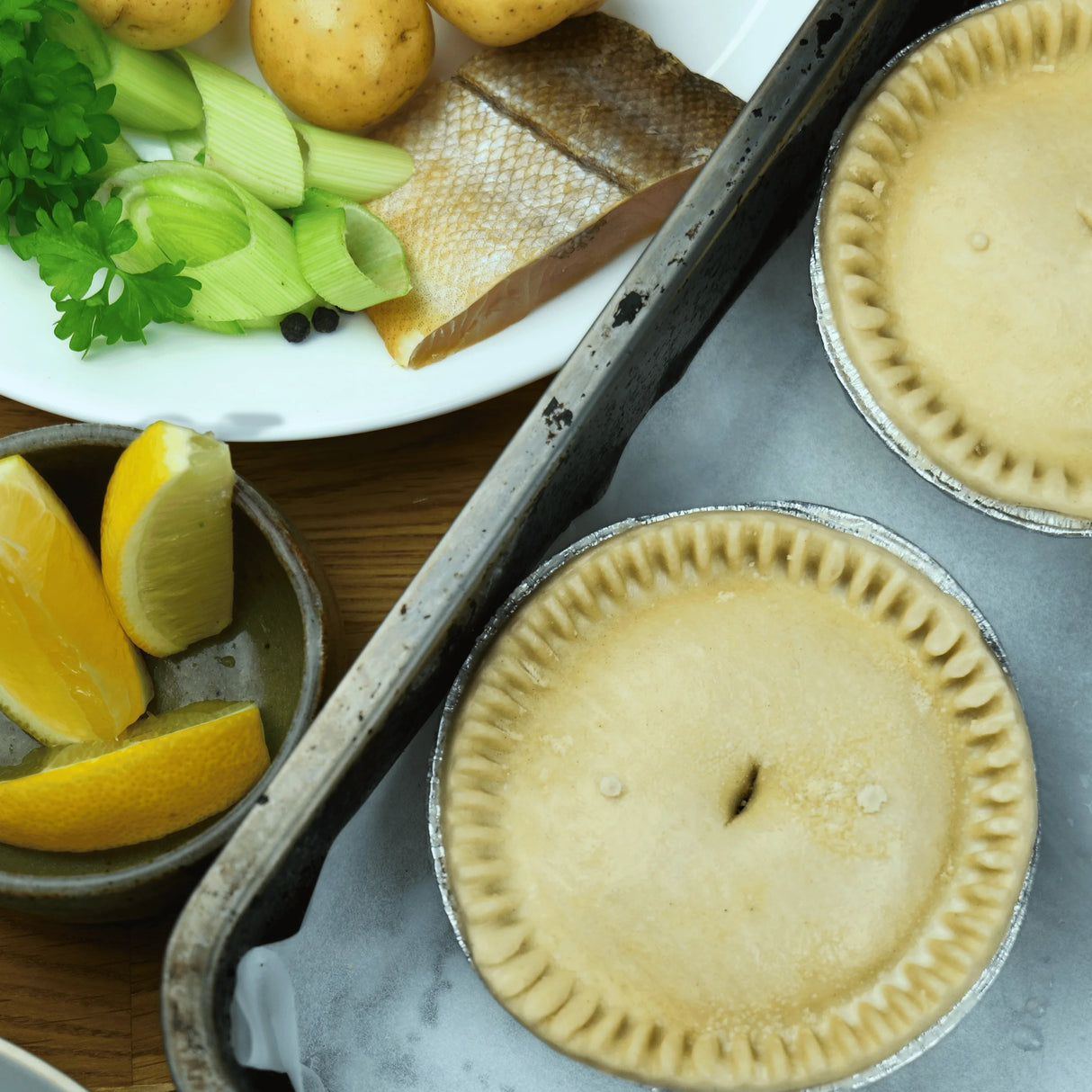 Unbaked Cornish fish pies in foil tins with fresh fish, potatoes, leek, parsley, and lemon wedges