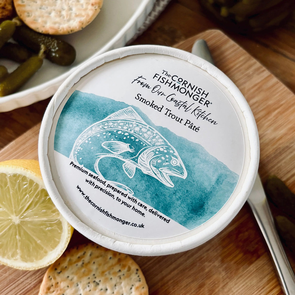 Tub of smoked trout pâté on a wooden board with crackers, lemon, and pickles