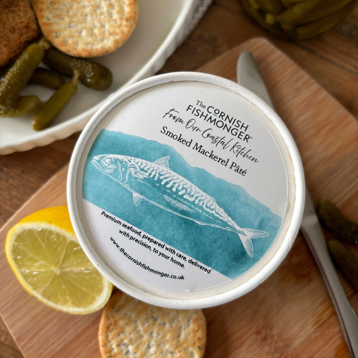 Smoked mackerel pâté in tub with lemon, crackers, and pickles on a wooden board