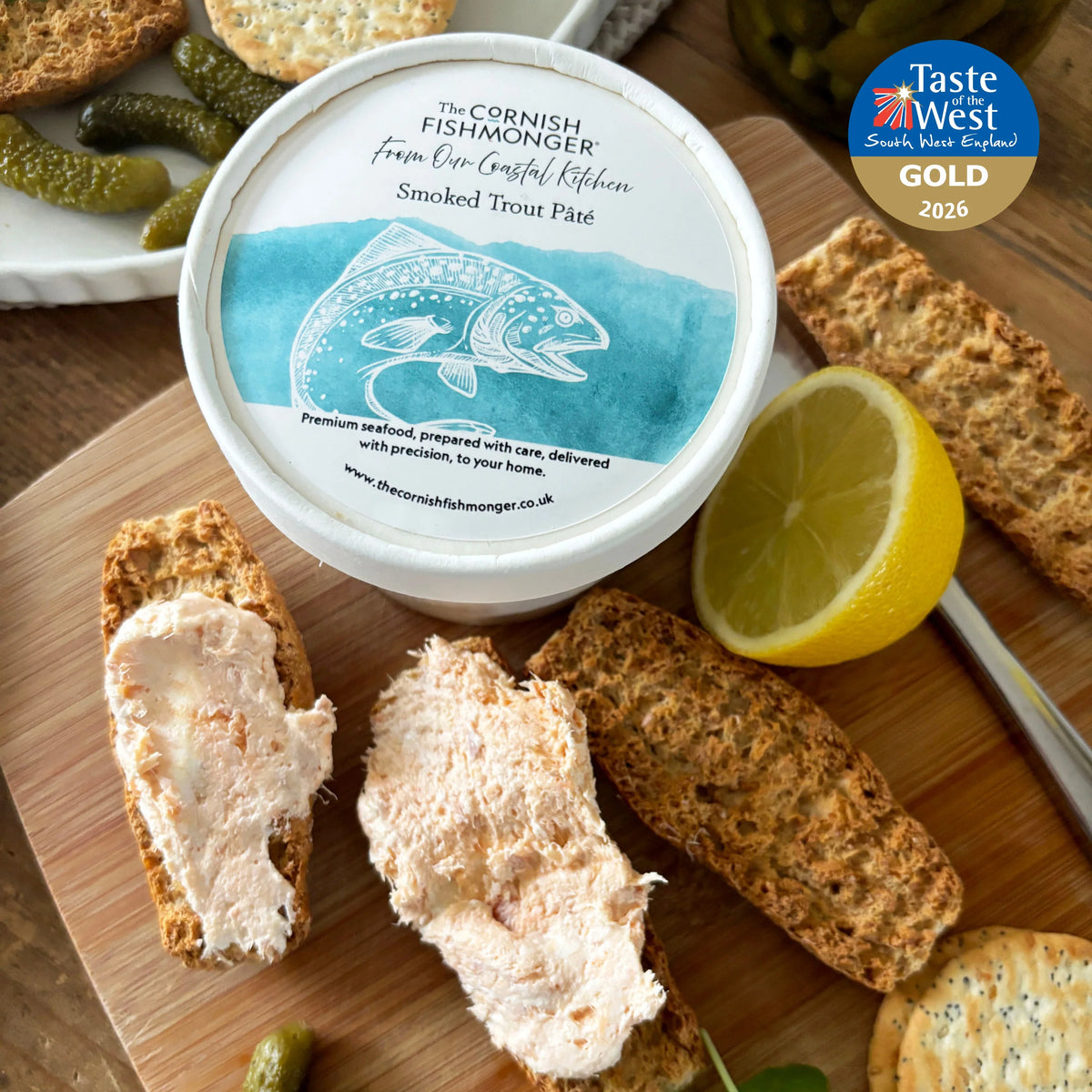 The Cornish Fishmonger Smoked Trout Pâté with crackers, lemon, and gherkins on a wooden board.