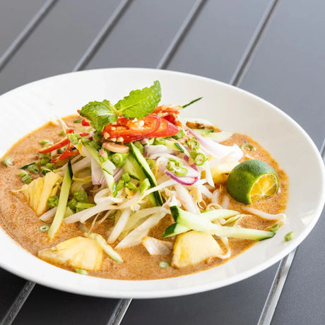 Fresh seafood laksa with sliced vegetables, pineapple, lime, and herbs in a white bowl