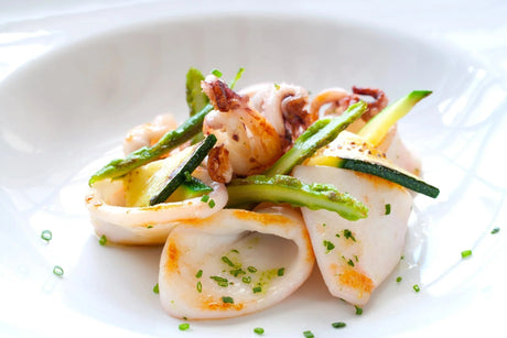 Grilled calamari with asparagus and zucchini on a white plate, garnished with herbs.