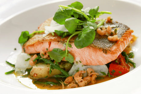 Seared salmon fillet with new potatoes, prawns, greens, and fresh herbs on a white plate