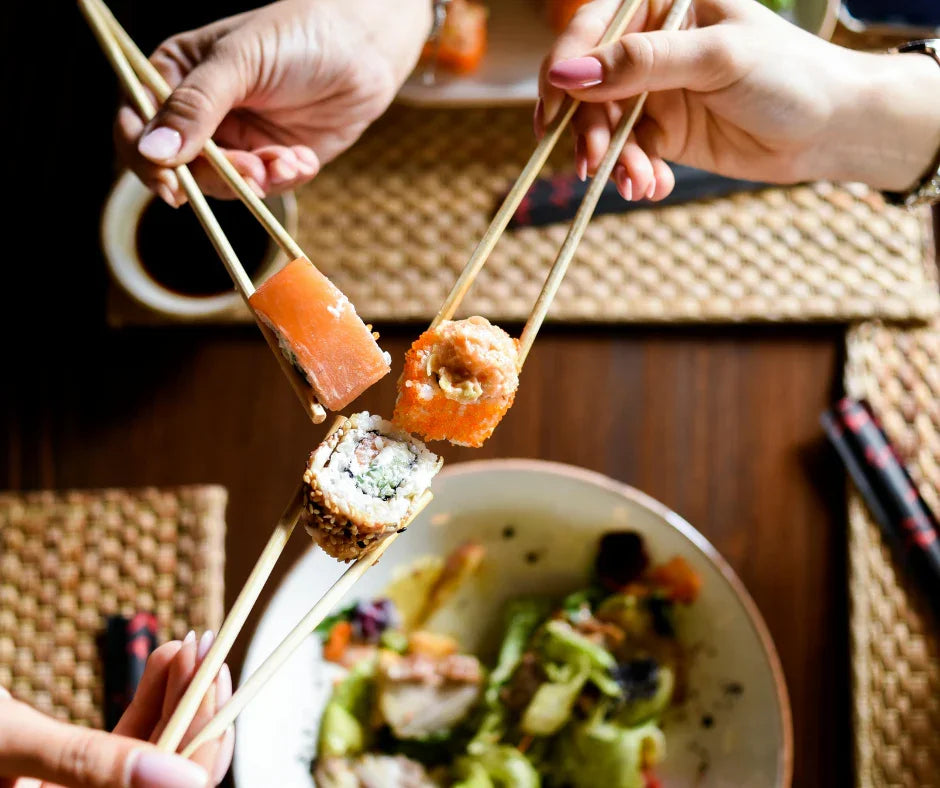 Three hands holding assorted sushi pieces with chopsticks over a table setting