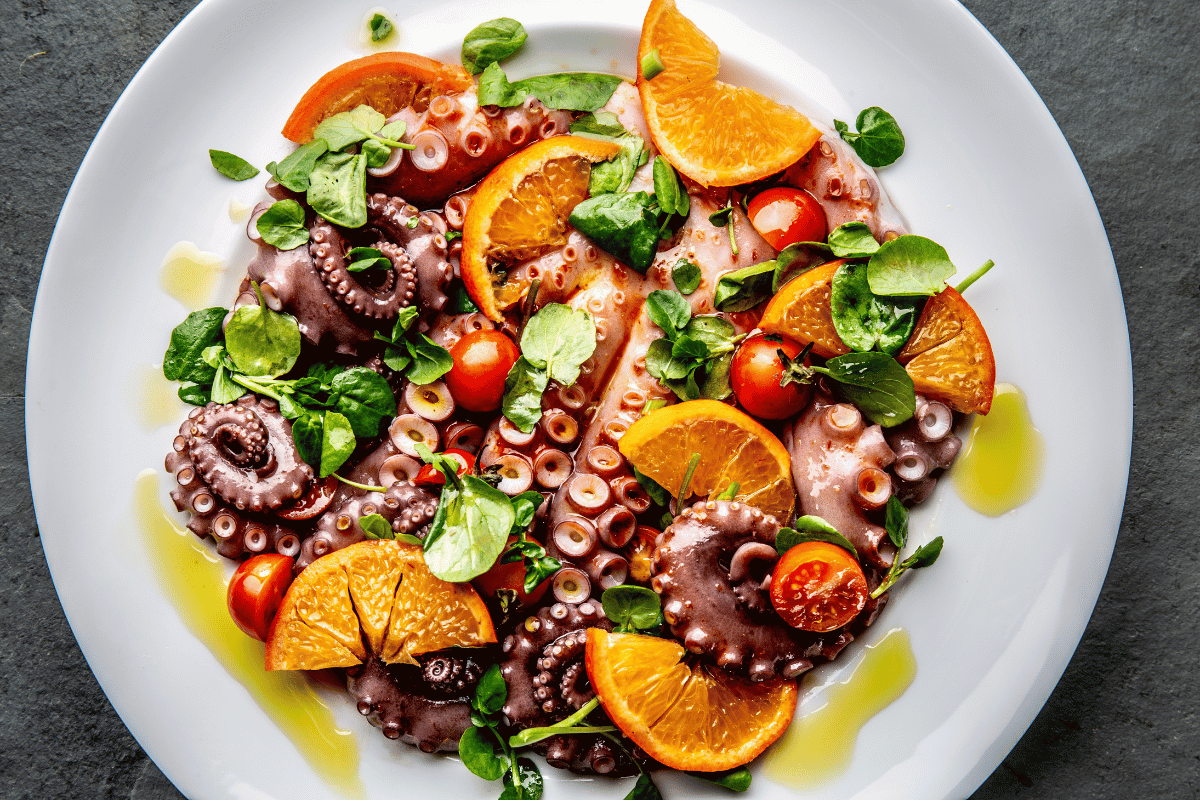 Octopus salad with orange slices, cherry tomatoes, fresh greens, and olive oil on a white plate