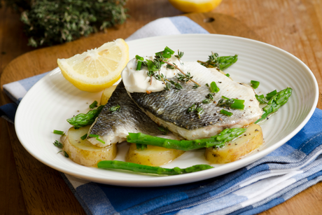 Pan-fried sea bass fillet with potatoes, asparagus, fresh herbs, and a lemon wedge on a white plate