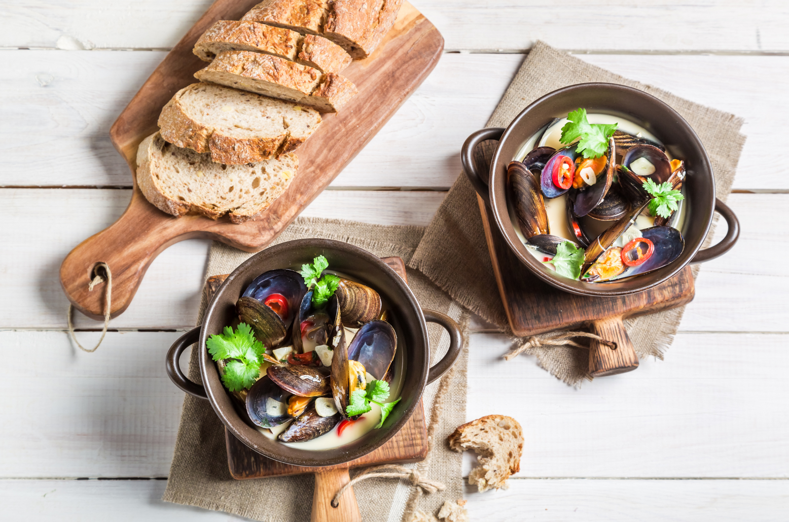Fresh cooked mussels in broth with sliced bread on a wooden table, garnished with herbs and chili